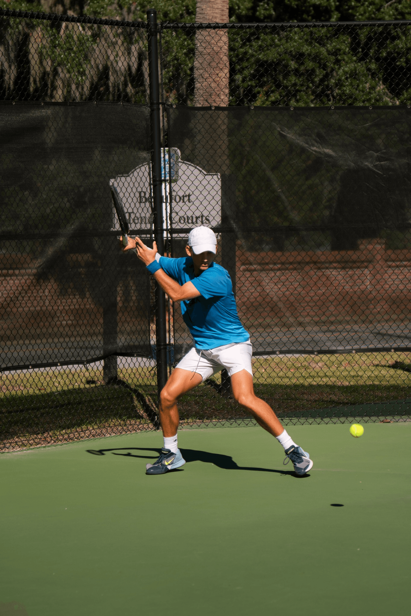 A player in a blue shirt and white cap setting up a forehand on a green hard court