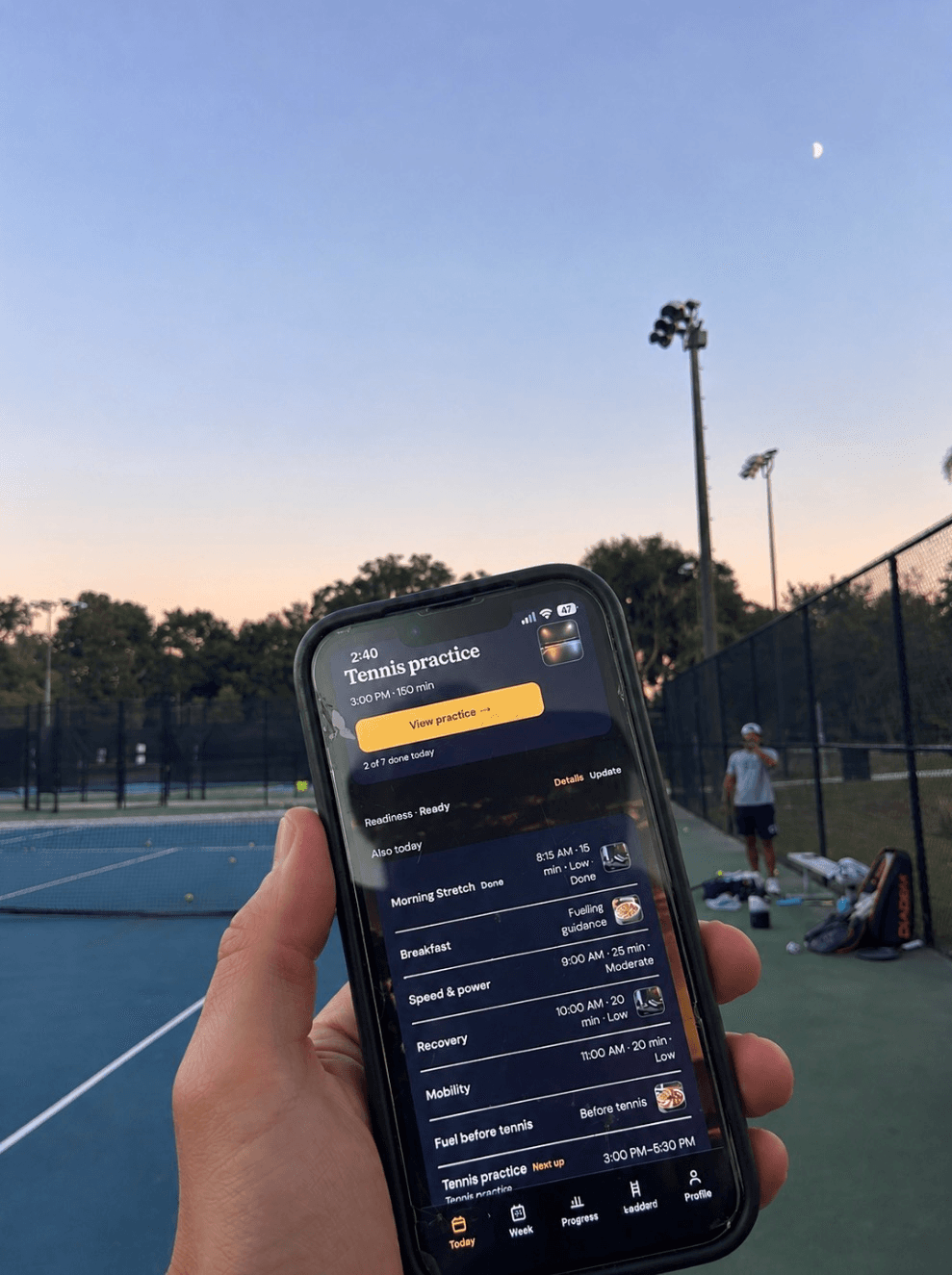 A hand holding a phone open to Acemore’s Today screen beside a tennis court at dusk